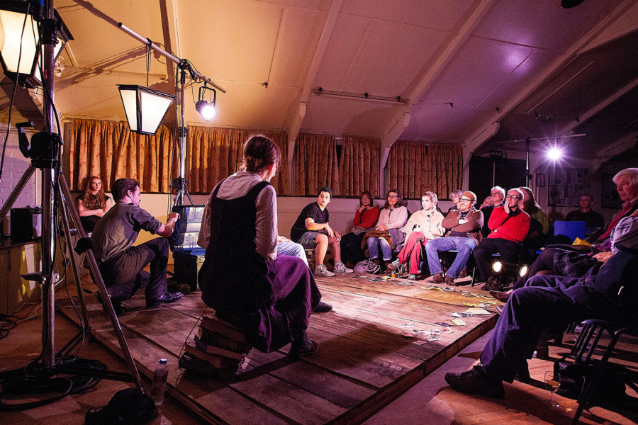 Group of people sat around a small wooden stage. There are some people sat on the stage with ornate looking lights.