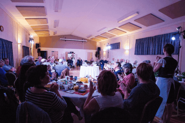 round tables of people at an event, there is somebody stood at the back speaking wearing an apron