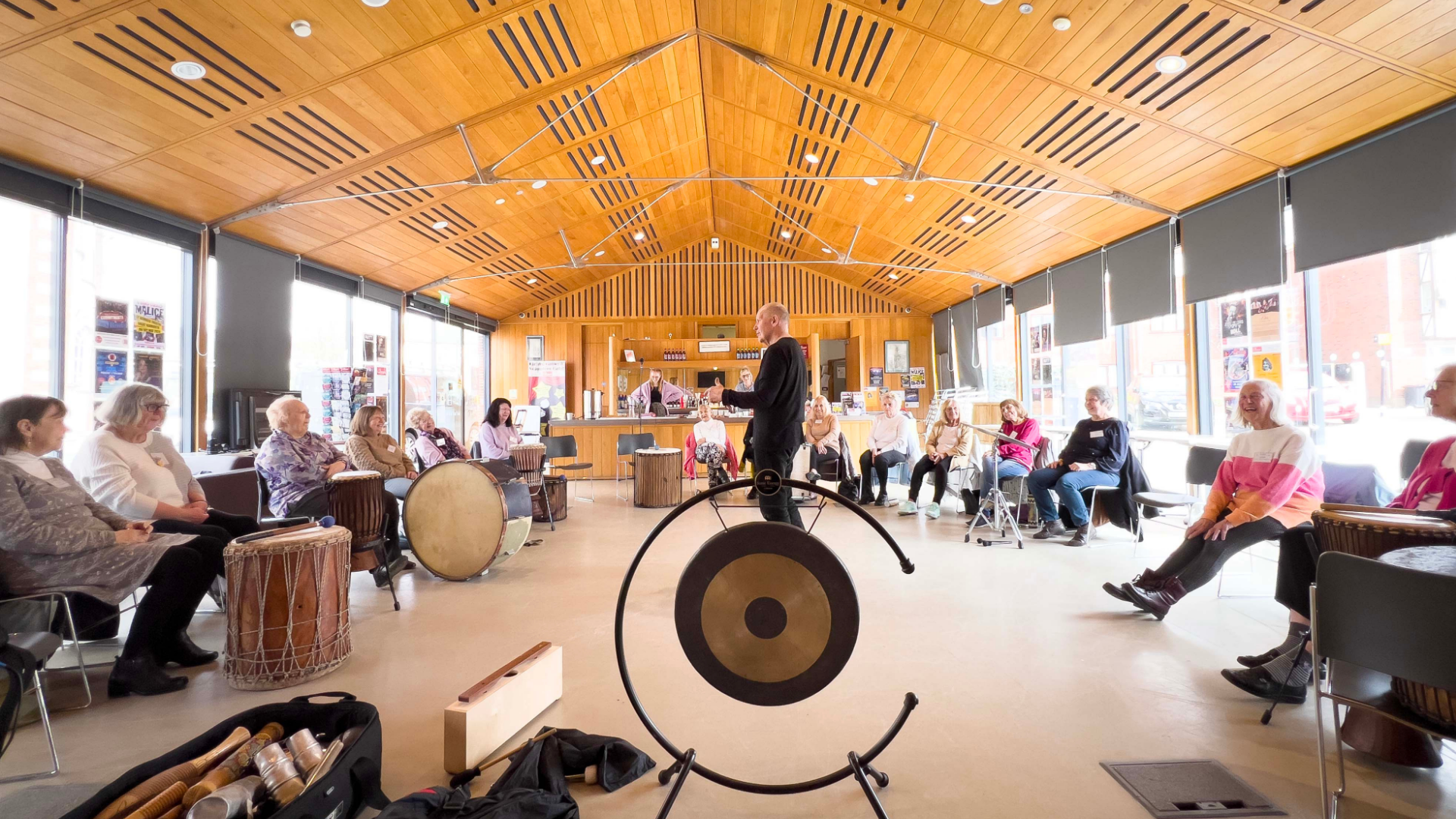 Group of people sat around in a large room with a wooden ceiling, they are all holding instruments and looking toward the leader in the middle. There is a a gong closest to the camera.