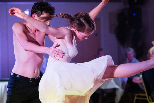 Two people on stage dancing. The person at the front is kicking their leg in the air while the person at the back is holding them steady.