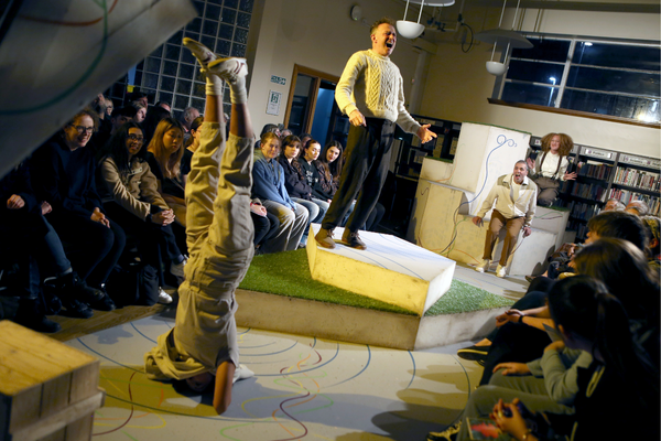 A long thin stage with audiences either side. The cast are performing.